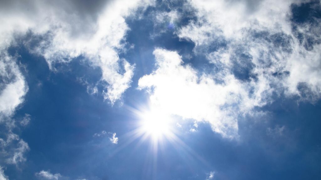 A vivid view of the sun shining through fluffy clouds in a bright blue sky.